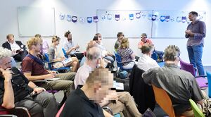Foto van de themasessie. Zichtbaar zijn ca 16 mensen die in verschillende luisterhoudingen in stoelen zitten, met daarvoor Gijsbert Kruithof, de trekker van deze sessie. Om privacyredenen zijn de gezichten onherkenbaar gemaakt. Op de achtergrond hangen vlaggetjes van de NORA Gebruikersdag.